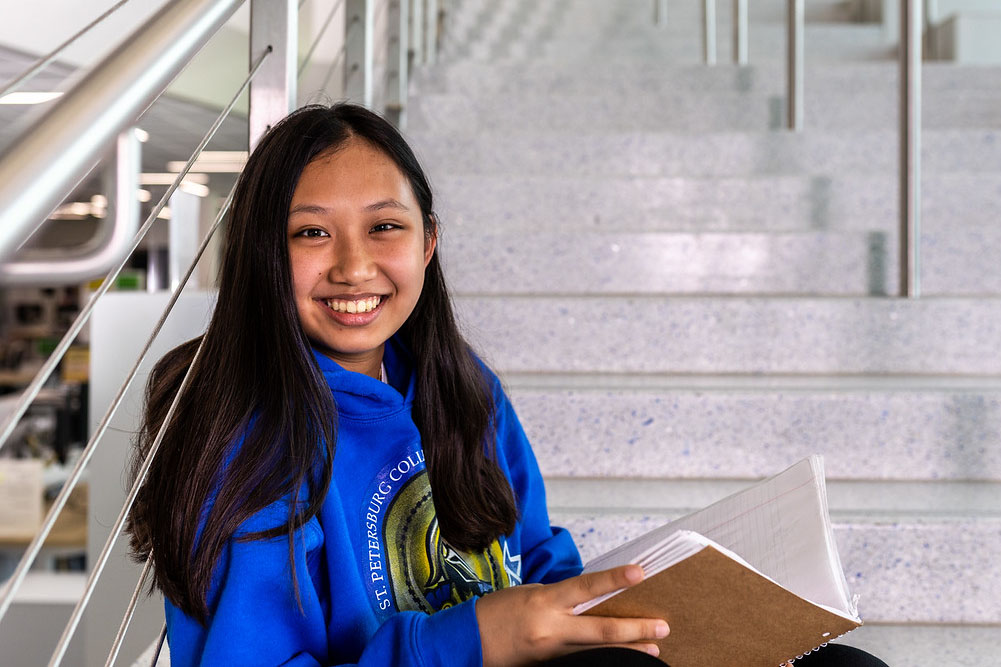 SPC student female student wearing an SPC sweatshit opening a brown notebook sitting on the stairs inside the Student Success Center on the St. Petersburg Gibbs Campus