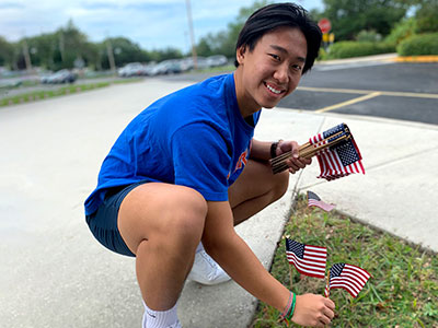 A male SPC student, wearing a ble shirt and shorts, planting small United States of America flags around campus.
