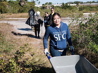 A female SPC student, wearing a blue SPC hoodie, pushing a wheel barrow as she volunteers for a cleanup event. 4 other students are pictured at a distance in the background.