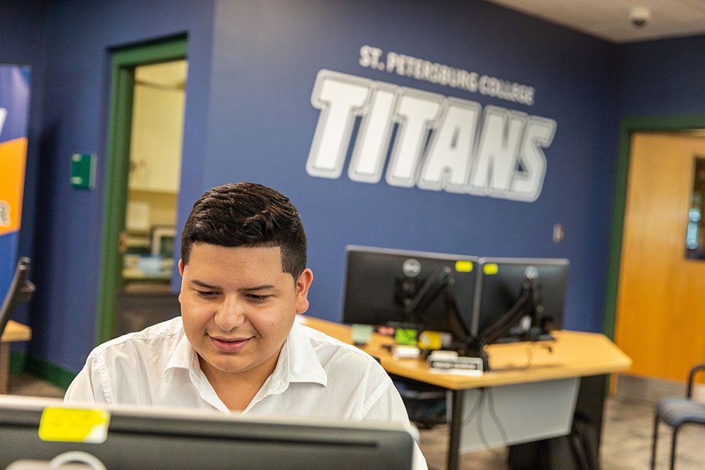 male SPC student at desk male student sitting at a computer in front of wall with Titans logo