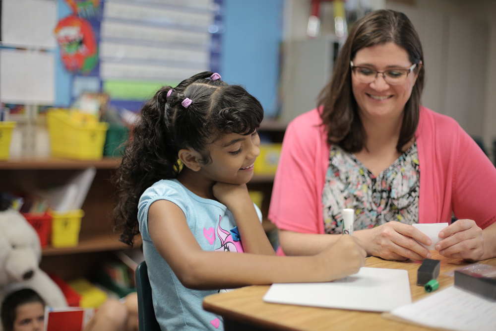 female teacher sitting with a student at a desk reading a book