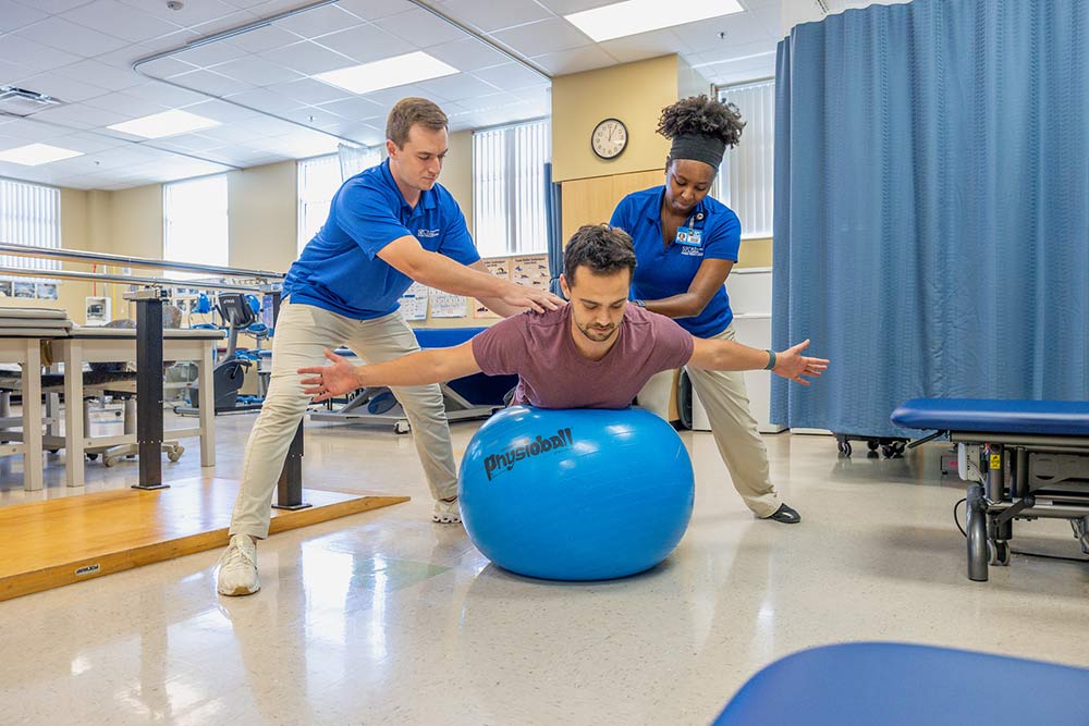 two physical therapy students helping a patient balance on a blue therapy ball