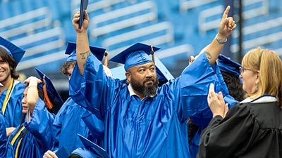 a graduate wearing a blue cap and gown holds his fingers up in a victory sign during commencement