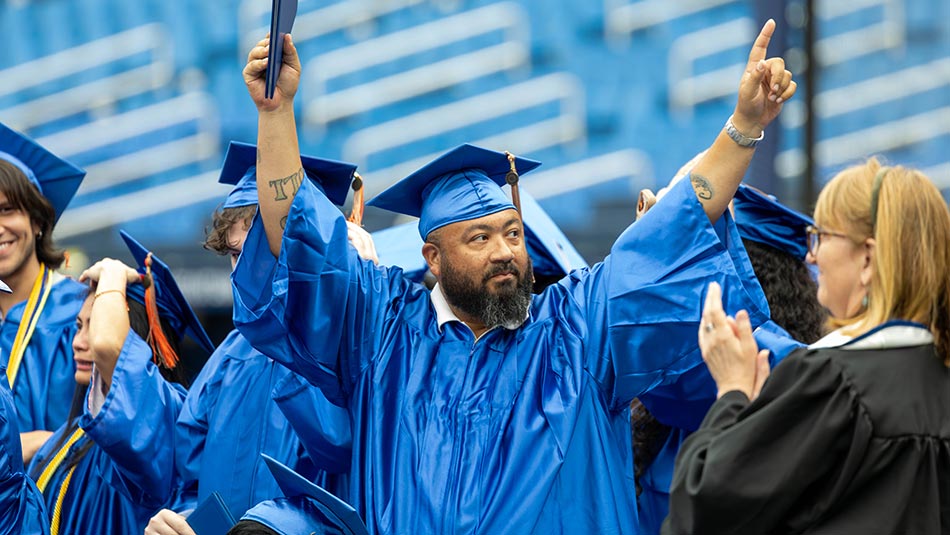 a graduate wearing a blue cap and gown holds his fingers up in a victory sign during commencement