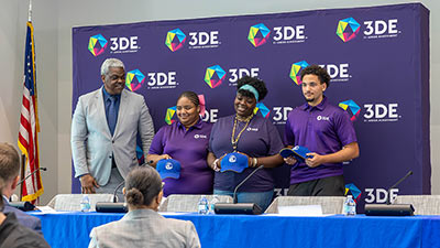 a group of 3DE participants standing on a stage smiling holding baseball caps