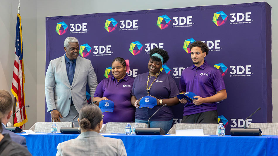 a group of 3DE participants standing on a stage smiling holding baseball caps