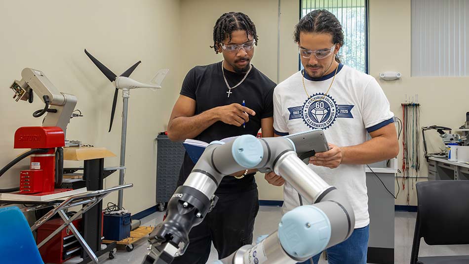 Two male students wearing a black shirt and white shirt with the words SPC wearing goggles in a manufacturing room