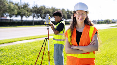 a male student using surveying equipment behind a female student wearing a hard hat and safety vest
