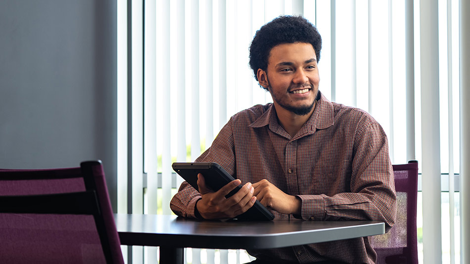 male student with a beard and brown shirt holding a tablet at a table