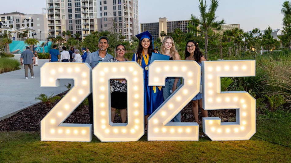 female graduate in her blue cap and gown celebrating wtih friends and family in front of a giant lit sign that says 2025