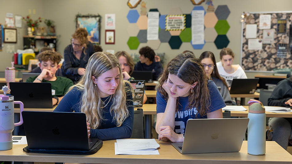 two female students studying together at a desk with the classroom in the background