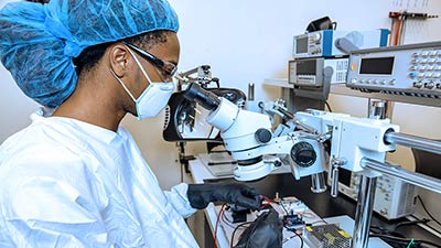 student in scrubs working at a microscope with black gloves on