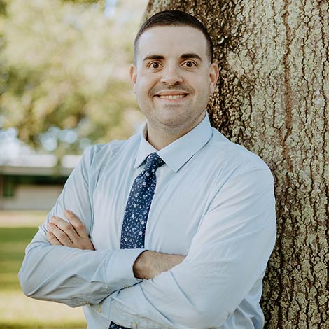 portrait of Eleftherios TJ Venieris wearing a blue tie, white button down shirt, leaning against a tree