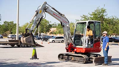 male student operating a heavy equipment excavator with a supervisor in a hard hat looking on