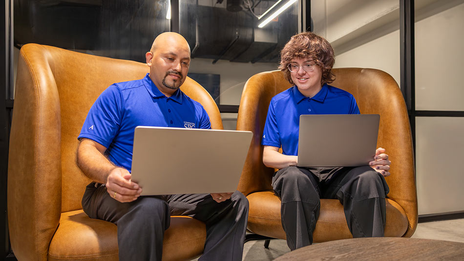 two male students wearing blue SPC shirts sitting together in brown leather chairs with laptops
