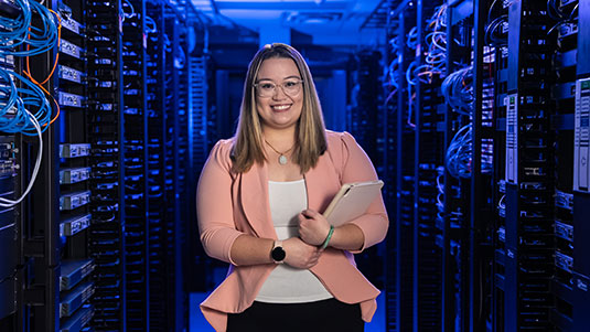 A female student, wearing a pink blazer and white top, poses in a dimly-lit server room