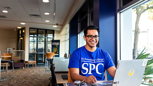A male student, wearing a blue SPC t-shirt, seated at a table inside a campus building, working at his laptop