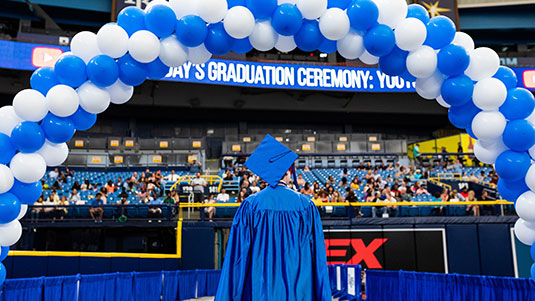 A graduate, wearing a blue cap and gown, walks away from the camera under an arch of blue and white balloons at the graduation ceremony
