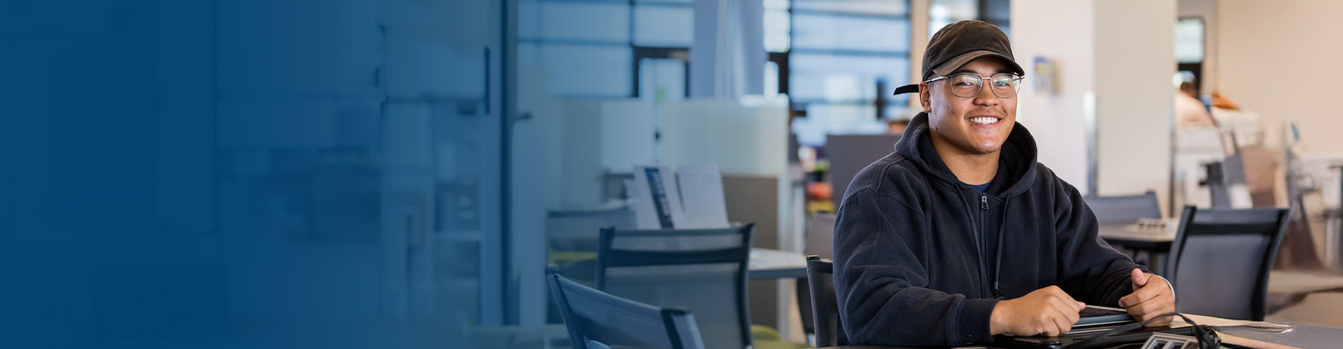 blue gradient image that fades into a smiling male student wearing glasses, a baseball cap and holding a pencil