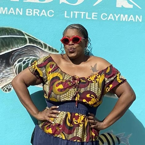 portrait of Kateka-Riggins wearing a brown patterned shirt in front of a blue backdrop