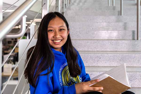 A female SPC student, wearing a blue SPC hoodie, sitting on the steps holding her notebook.