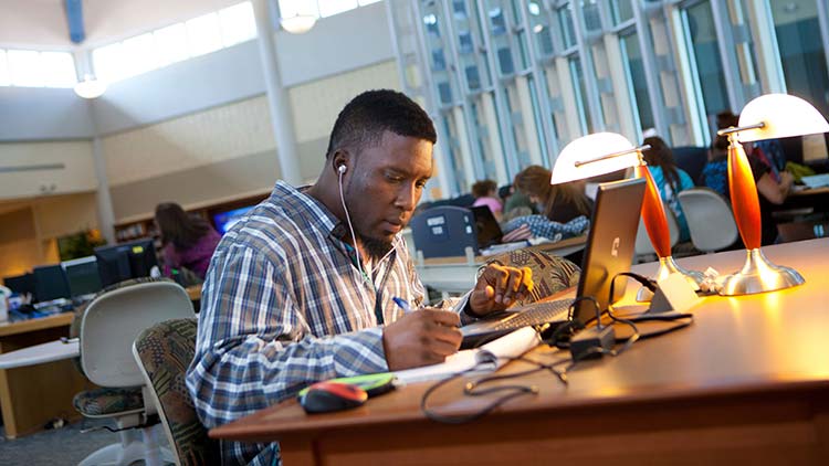 A male student, sitting at his laptop in a large room, writing on a notepad.