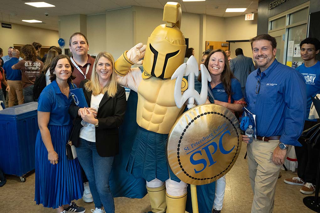 SPC Day 2024 Five SPC staff posing with Titus the Titan at SPC Day 2024, with many people conversing in the background.