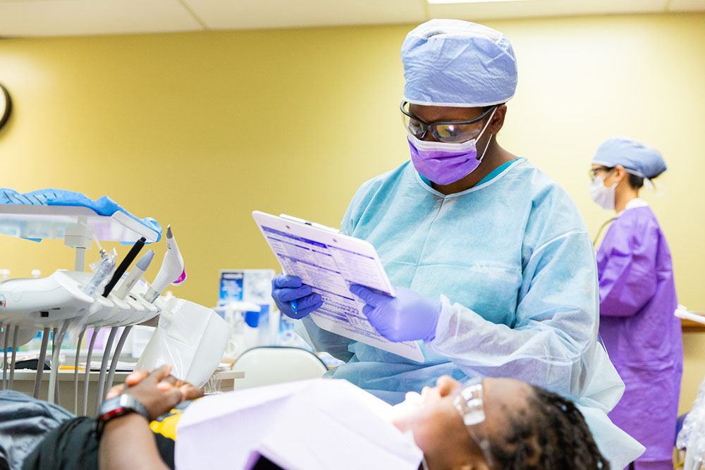 female dental hygiene student wearing a mask and holding a clipboard in front of a seated patient