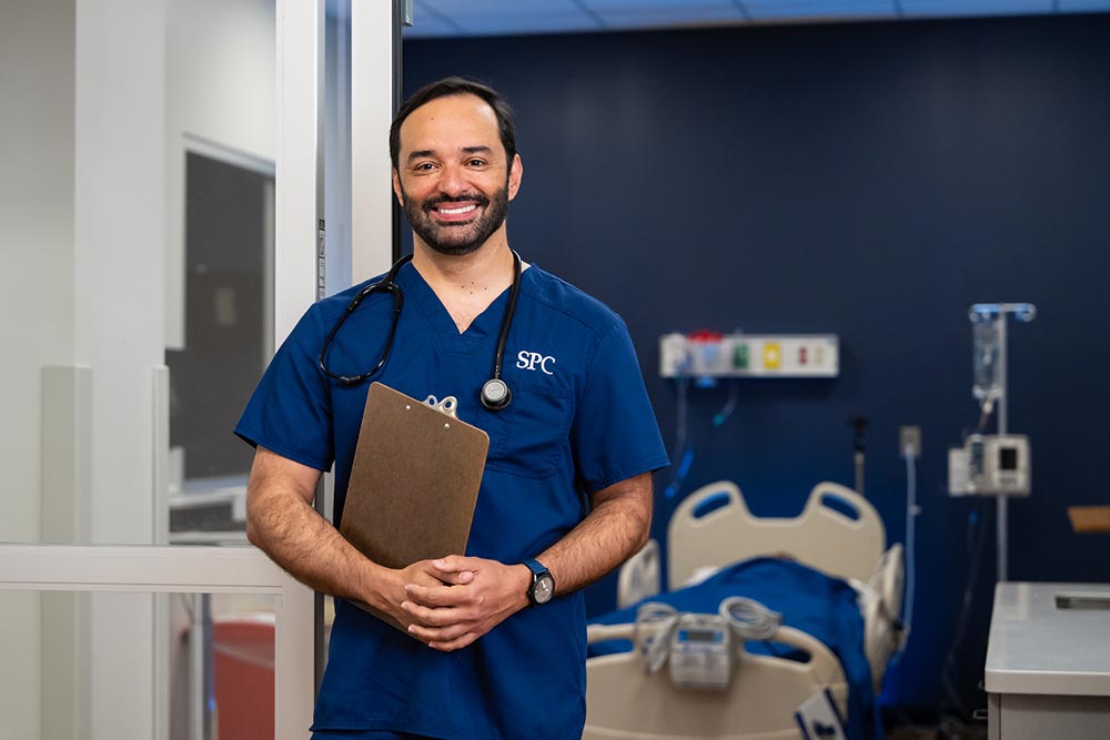 SPC student male student dressed in blue scrubs holding a clipboard in front of a hospital room