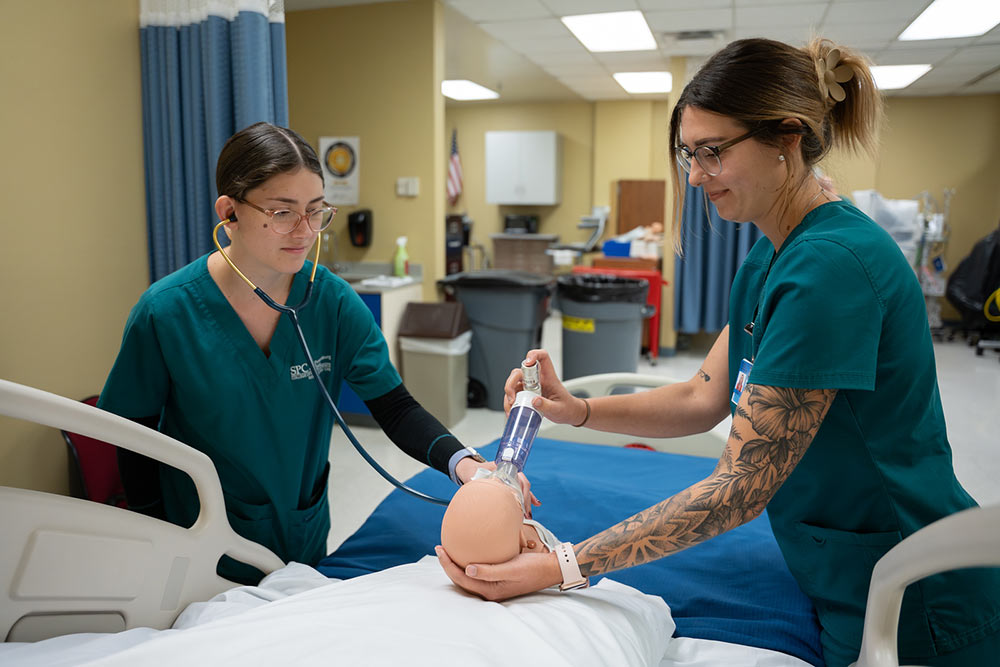 two female health students in green scrubs putting a respiratory device on a model baby