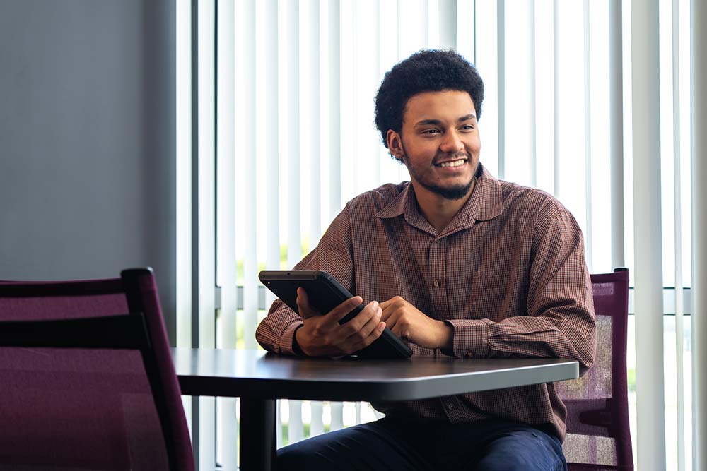 male student holding a tablet wearing a brown button down shirt sitting at a table