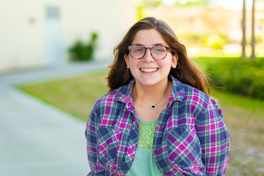 female student wearing glasses, a checked flannel shirt over a green shirt and a heart shaped necklace