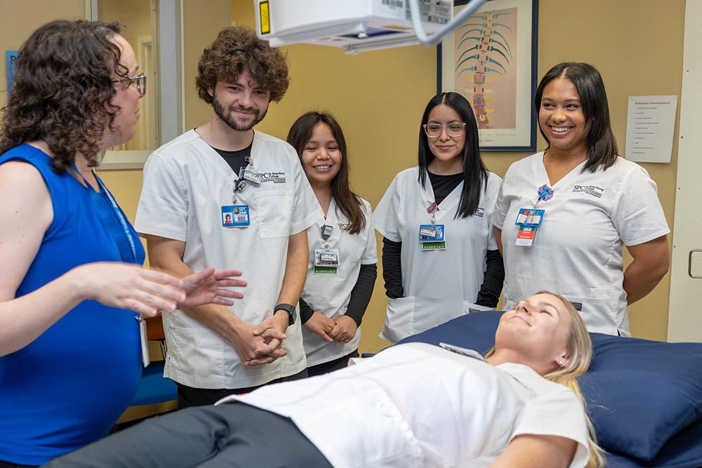 group of radiography students standing around a table listening to a lecture from a female professor