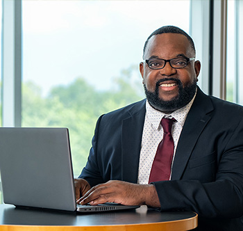 man in a black suit sitting at a laptop in front of a window