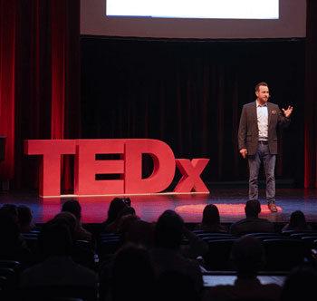 Man presents on a stage. He's flanked by a large 3D logo signs to the left of him of the TEDx logo