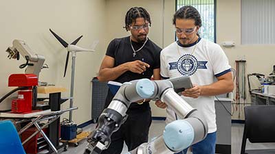 Two male students wearing a black shirt and white shirt with the words SPC wearing goggles in a manufacturing room