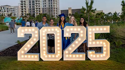 female graduate in her blue cap and gown celebrating wtih friends and family in front of a giant lit sign that says 2025