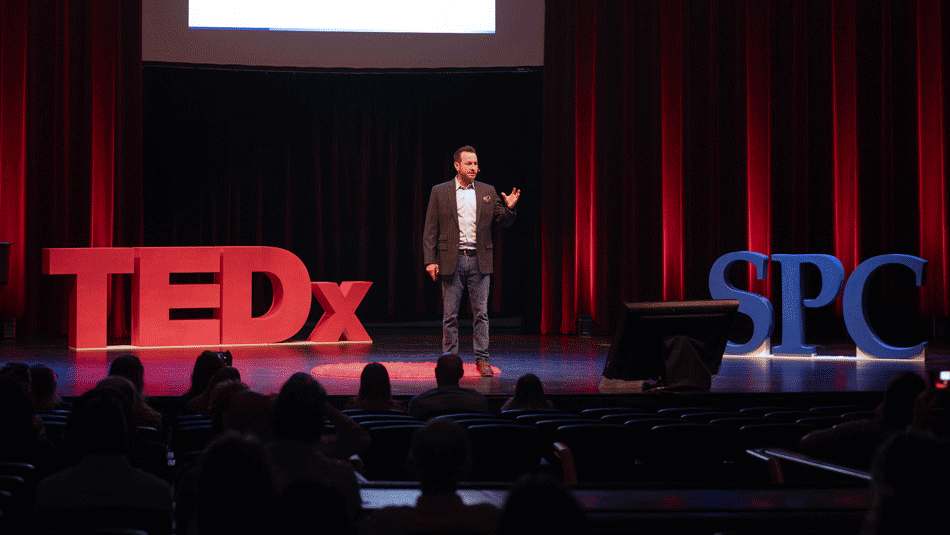 a male presenter on stage between 3-d modesl with the letters TEDx and SPC