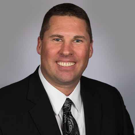 Stephen Andrasik posing in a black suit and black tie, in front of a gray background.