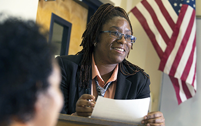 female teacher in front of a class wearing a black jacket in front of an american flag