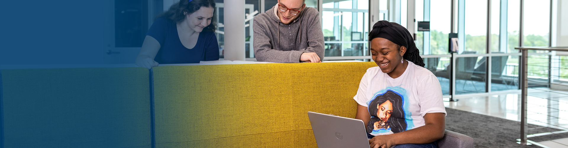 blue gradient image that fades into a female student sitting on a yellow couch laughing with a laptop in her lap and two students standing behind her laughing