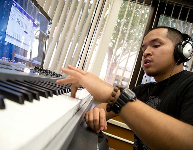 A male student, wearing headphones, playing an electronic keyboard while taking a quiz on music fundamentals from a computer screen.