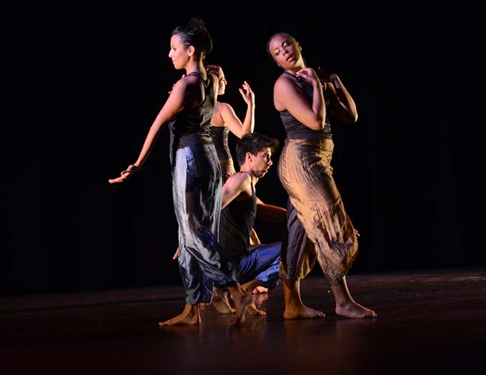 Four dancers, three female and one male, under the spotlight on a dark stage.